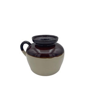 Op de afbeelding: Een keramische boterpot met een tweekleurig ontwerp, voorzien van een glanzend donkerbruin geglazuurd bovendeel en een gladde, crèmekleurige basis. De pot heeft een enkel gebogen handvat aan de zijkant en een brede opening met een platte rand.