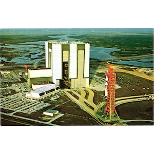 May include: Vintage postcard featuring a large white and green building with a tall red and orange launch structure. The scene is set against a backdrop of water and green land, with a parking lot and roads in the foreground. The postcard has a retro aesthetic.