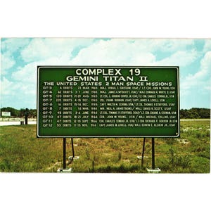 Puede incluir: Una postal vintage con un letrero verde para las misiones espaciales Complex 19 Gemini Titan II. El letrero enumera los detalles de la misión, incluidas las fechas, las órbitas y los miembros de la tripulación. El fondo muestra una zona de césped y un cielo parcialmente nublado.