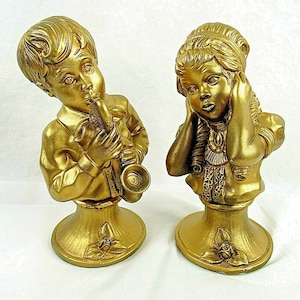 Puede incluir: Dos figuritas doradas, una tocando una trompeta y la otra con las manos sobre las orejas, sobre bases decorativas.