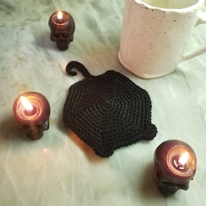 Pode incluir: Tapete de crochê preto em forma de morcego com um centro redondo. O tapete é cercado por três pequenos porta-velas de caveira preta com velas acesas.