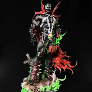 Puede incluir: Figura de acción detallada de Spawn, un personaje de cómic, de pie sobre una base rocosa. La figura es predominantemente negra con acentos blancos, una capa roja y efectos de energía verde. Cuenta con cadenas, un cinturón de calavera y detalles puntiagudos.