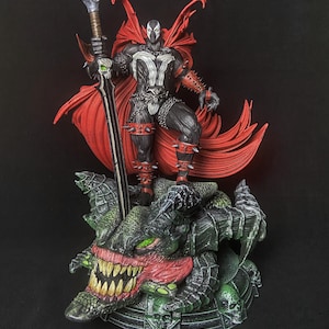 Puede incluir: Figura de acción de Spawn sobre una base detallada con una cabeza de monstruo. La figura es negra y blanca con una capa roja y detalles puntiagudos, sosteniendo una gran espada. La base es verde y roja.