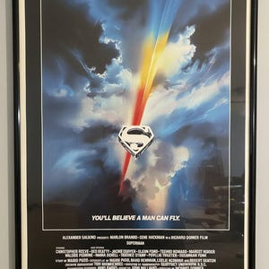 May include: A framed Superman movie poster featuring a central silver S-shield emblem against a dark blue sky with bright light streaks and clouds. The text reads: YOU'LL BELIEVE A MAN CAN FLY, along with cast and production credits in white lettering.