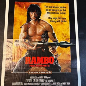 May include: A Rambo: First Blood Part II film poster featuring Sylvester Stallone holding a rocket launcher against a fiery orange background. The text reads: They sent him on a mission and set him up to fail. But they made one mistake. They forgot they were dealing with Rambo. No man, no law, no war can stop him.