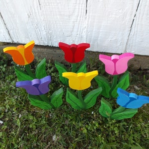 May include: A set of seven wooden tulip flower decorations painted in various colors, including red, pink, yellow, orange, blue, and purple. The tulips are arranged in a row on a bed of green grass.