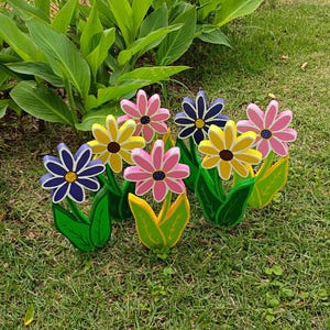 May include: A cluster of colorful wooden flowers with green leaves, set in grass. The flowers are painted in shades of pink, yellow, and blue, with brown centers. The leaves are green and yellow. A decorative garden accent.