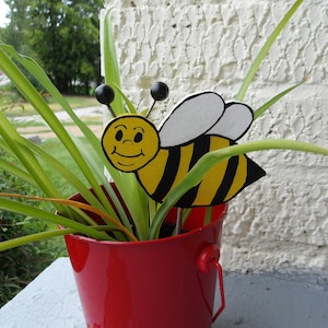 May include: A red bucket planter with a green plant and a yellow and black bee decoration with a smiling face.