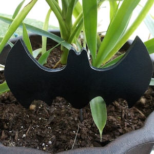 May include: Black bat-shaped garden stake, inserted into the soil of a potted plant. The plant has long, green leaves.