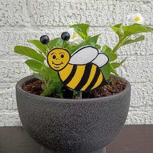 May include: A whimsical, hand-painted wooden bee garden stake with a smiling face, yellow and black stripes, and white wings. The bee is placed in a dark gray ceramic pot with green foliage and white flowers. The background is a white brick wall.