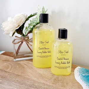 May include: Two clear plastic bottles of bath products with black lids and labels that read "Cottage Fresh Scents Coastal Breeze Luxury Bubble Bath". The bottles are on a wooden surface with a white and blue background.