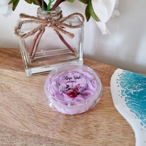 May include: A small clear container with a purple whipped soap inside. The container has a label that says "Cottage Fresh Scents" with a floral design.