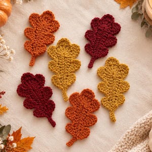May include: A set of six crocheted oak leaf coasters in autumn colors, including two deep burgundy, two mustard yellow, and two burnt orange leaves. Each leaf features a textured, hand-stitched design with a central stem and lobed edges.
