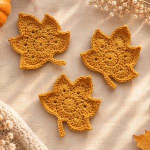 May include: Three mustard yellow crochet maple leaf coasters. Each leaf features a detailed, textured stitch pattern with a central stem and five distinct lobes. The coasters are arranged in a cluster, showcasing the intricate yarn work and the vibrant, warm autumn-inspired color of the thread used to create these decorative textile items.