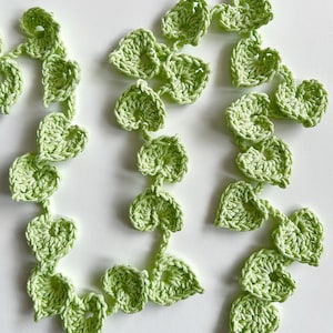 May include: Crocheted garland featuring a repeating pattern of green leaves. The leaves are a light green color and are connected by a thin, crocheted vine. The garland is likely handmade and suitable for home decor.