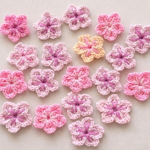 May include: A collection of small, five-petaled crochet flowers in various shades of pink, light purple, and one pale yellow. Each flower features a textured, stitched center and delicate yarn loops forming the petals, arranged in a scattered pattern.