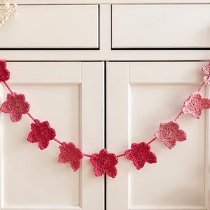 May include: A decorative garland featuring ten crocheted flower motifs in varying shades of pink, ranging from deep magenta to soft rose. The flowers are connected by a thin matching yarn string, creating a scalloped drape across the cabinet doors.