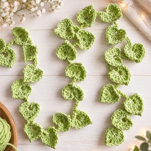 May include: A long, decorative garland made of light green crocheted yarn, featuring a series of interconnected heart-shaped leaves. The crochet work displays a consistent stitch pattern throughout the entire length of the vine-like strand.