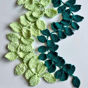 May include: Crocheted leaf garland in shades of green. The leaves are arranged in a flowing pattern, transitioning from light green to a deep teal. The handmade garland is likely used for home decor or crafting projects.