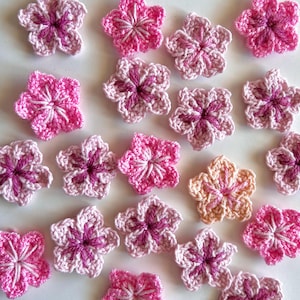Könnte beinhalten: Eine Ansammlung handgehäkelter Blumen in verschiedenen Rosa- und Weißtönen. Die Blumen haben fünf Blütenblätter und eine dunkelrosa Mitte, ideal für Bastelprojekte oder Verzierungen.