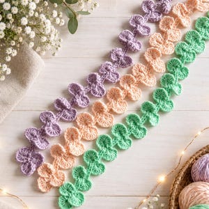 May include: Three long, decorative crochet trim strips arranged diagonally. Each strip features a repeating pattern of small, rounded, fan-like petals. The colors of the three strips are light purple, soft peach, and mint green.
