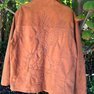 May include: A brown suede jacket with an embroidered eagle and snake design on the back. The jacket has a collar and a button closure. The jacket is decorated with a stitched barbed wire design around the edges.