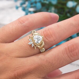 May include: A gold heart-shaped ring with a halo of smaller diamonds. The ring is set in a Celtic-style band with two hands clasping the heart.