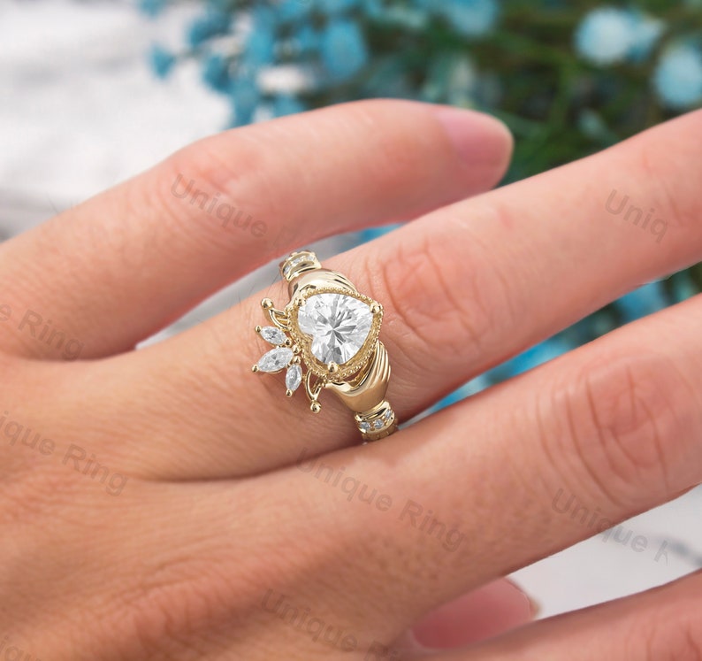 May include: A gold heart-shaped ring with a halo of smaller diamonds. The ring is set in a Celtic-style band with two hands clasping the heart.