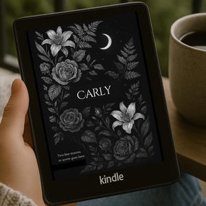 Può includere: Un e-reader Kindle nero con un design floreale con il nome "CARLY" sullo schermo. Il design presenta rose, gigli e una luna crescente. Il Kindle è tenuto in mano, con una tazza di caffè sullo sfondo.