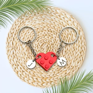 May include: A red heart-shaped keychain made of Lego bricks with the letters "J" and "M" on the sides. The keychain is attached to two silver key rings.