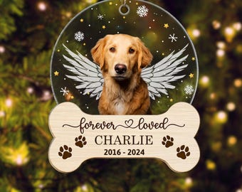 May include: A round, clear Christmas ornament featuring a golden retriever with angel wings. Below the dog is a wooden bone shape with the words "forever loved" and "CHARLIE 2016-2024" along with paw prints. The ornament hangs from a gold string.