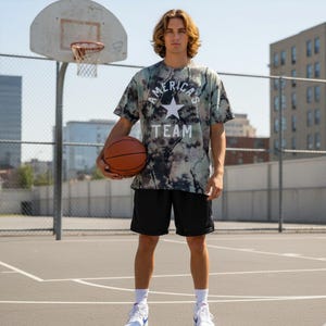 Pode incluir: Uma pessoa em uma quadra de basquete segura uma bola de basquete. Usa uma camiseta tie-dye com a frase "AMERICA'S TEAM" e uma estrela, calções pretos e tênis brancos. Uma cesta de basquete está ao fundo.
