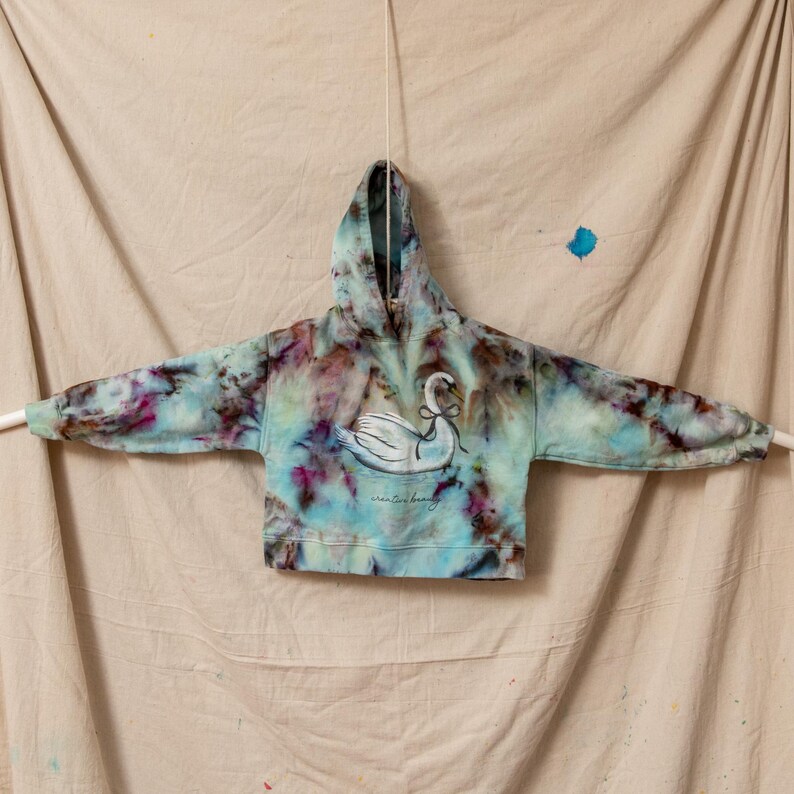 A tie-dye hoodie in shades of blue, purple, and brown. The hoodie features a white swan graphic with a bow and the words 'winter love beauty' printed below. The hoodie is hanging against a beige backdrop.