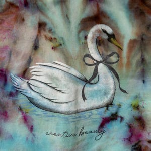 K&ouml;nnte beinhalten: Illustration eines wei&szlig;en Schwans mit schwarzer Schleife, der auf Wasser schwimmt. Der Hintergrund hat einen Aquarell-Effekt mit Blau-, Rosa- und Braunt&ouml;nen. Die Worte "creative beauty" sind unten in Kursivschrift geschrieben.