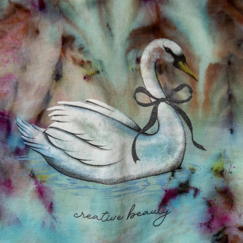 K&ouml;nnte beinhalten: Illustration eines wei&szlig;en Schwans mit schwarzer Schleife, der auf Wasser schwimmt. Der Hintergrund hat einen Aquarell-Effekt mit Blau-, Rosa- und Braunt&ouml;nen. Die Worte "creative beauty" sind unten in Kursivschrift geschrieben.