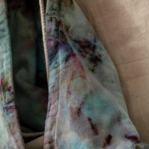 K&ouml;nnte beinhalten: Nahaufnahme eines Stoffes mit Batikmuster in verschiedenen Farben, darunter Blau, Rosa und Braun. Der Stoff scheint aus einem weichen Material, m&ouml;glicherweise Baumwolle, zu bestehen. Die Kanten sind sichtbar, mit einem Kordelzug.