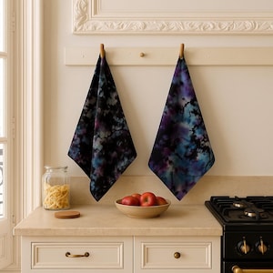 May include: Two tie-dye kitchen towels with a black, purple, and teal pattern hang from a wooden rack. The towels are triangular and have a marbled appearance. A bowl of red apples sits on the counter, along with a glass jar and a wooden coaster.