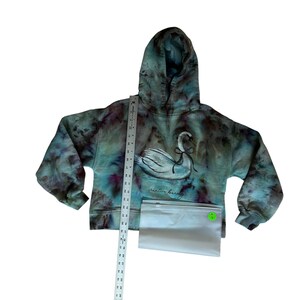 K&ouml;nnte beinhalten: Ein Kapuzenpullover mit Batikmuster und einem Schwan-Motiv. Der Pullover hat eine Mischung aus teal, lila und braunen Farben. Der Schwan ist wei&szlig; mit einer schwarzen Schleife. Der Text "creative beauty" steht unter dem Schwan. Ein Lineal und eine transparente T&uuml;te sind ebenfalls im Bild.