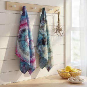 May include: Two tie-dye towels hang on a wooden rack against a white wall. One towel features pink, blue, and gray swirls, while the other has blue and gray patterns. A basket of lemons and a small dish are on the wooden counter.