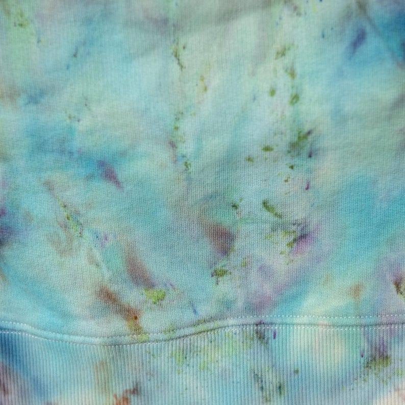 K&ouml;nnte beinhalten: Nahaufnahme eines Stoffes im Batik-Stil in Blau-, Gr&uuml;n- und Lilat&ouml;nen. Der Stoff hat unten eine gerippte Textur und insgesamt ein weiches, verschwommenes Aussehen. Die Farben sind gemischt und erzeugen einen Aquarelleffekt.