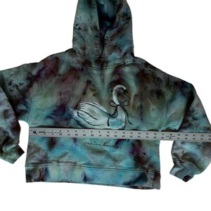 K&ouml;nnte beinhalten: Ein Kapuzenpullover mit Batikmuster in T&uuml;rkis, Lila und Wei&szlig;, mit einer Schwan-Illustration und einer schwarzen Schleife. Der Text "creative beauty" ist darunter aufgedruckt. Der Hoodie hat eine Kapuze und lange &Auml;rmel.