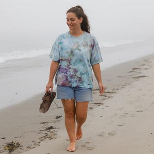 May include: A person wearing a blue tie-dye t-shirt and denim shorts walks barefoot on a sandy beach, holding brown sandals. The t-shirt has a unique pattern with shades of blue, purple, and green. The background is a foggy beach scene.