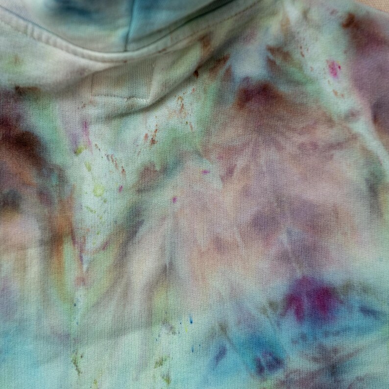 K&ouml;nnte beinhalten: Nahaufnahme eines Kapuzenpullovers mit Batikmuster in Blau-, Lila-, Gr&uuml;n- und Braunt&ouml;nen. Der Stoff hat ein weiches, strukturiertes Aussehen mit einem dezenten Muster. Die Kapuze ist oben sichtbar, mit einem Kordelzug.