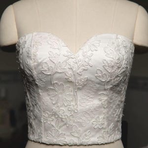 Puede incluir: Corsé blanco sin tirantes con superposición de encaje floral. El bustier se muestra en un maniquí y presenta un escote corazón. La prenda está adornada con delicados patrones florales bordados. El número "82" está impreso en el maniquí.
