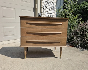 May include: A mid-century modern dresser with three drawers and brass legs. The dresser is made of wood and has a light brown finish.