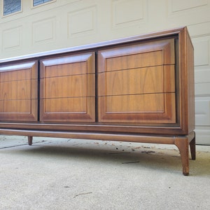 May include: A mid-century modern dresser with six drawers and a wood finish. The dresser has a simple design with clean lines and a slightly curved base.