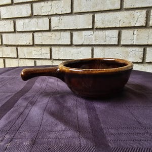 Puede incluir: Un cuenco de cerámica marrón con una sola asa. El cuenco tiene un acabado esmaltado y está sobre un mantel morado.