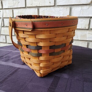 May include: A woven basket with a brown, green and red trim. The basket has a handle and is made of natural wood.