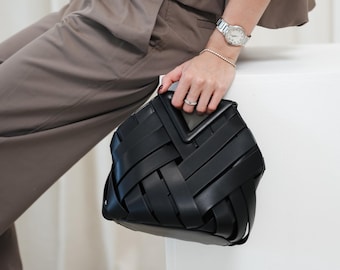 May include: A black woven leather handbag with a geometric design. The bag is held by a person wearing a brown outfit and a silver watch. The bag's woven pattern creates a textured surface.