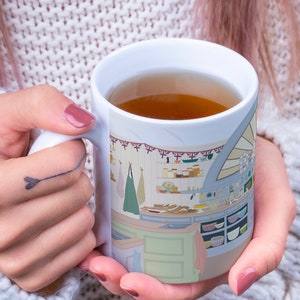May include: A white ceramic mug with a colorful illustration of a kitchen interior. The mug is filled with a brown liquid, possibly tea or coffee.